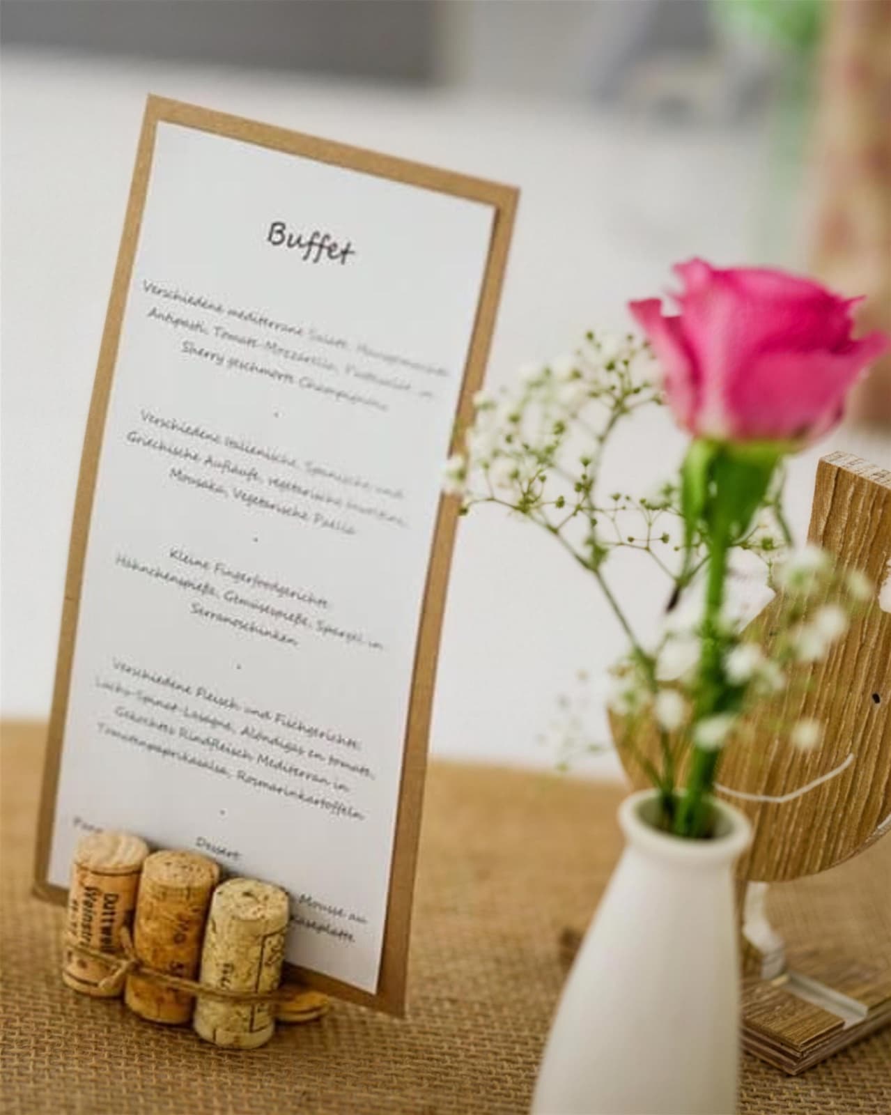 Handgeschriebene Buffetkarte in einem Halter aus Weinkorken auf einem Jutetischläufer, daneben eine pinke Rose mit Schleierkraut in einer kleinen weißen Vase.