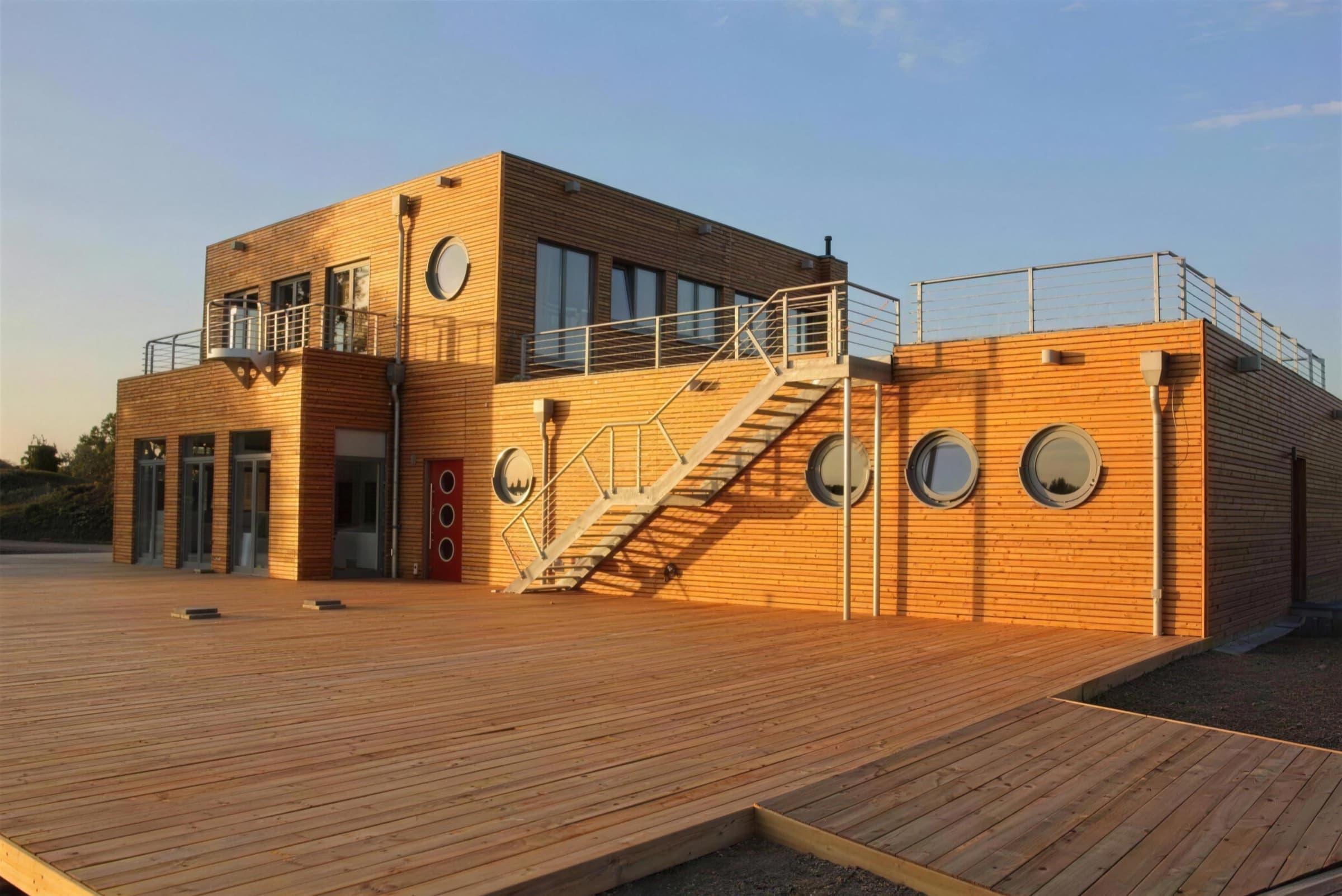 Zweigeschossiges Gebäude mit heller Holzfassade, runden Bullaugenfenstern und Außentreppe im warmen Abendlicht, davor eine weite Holzterrasse.