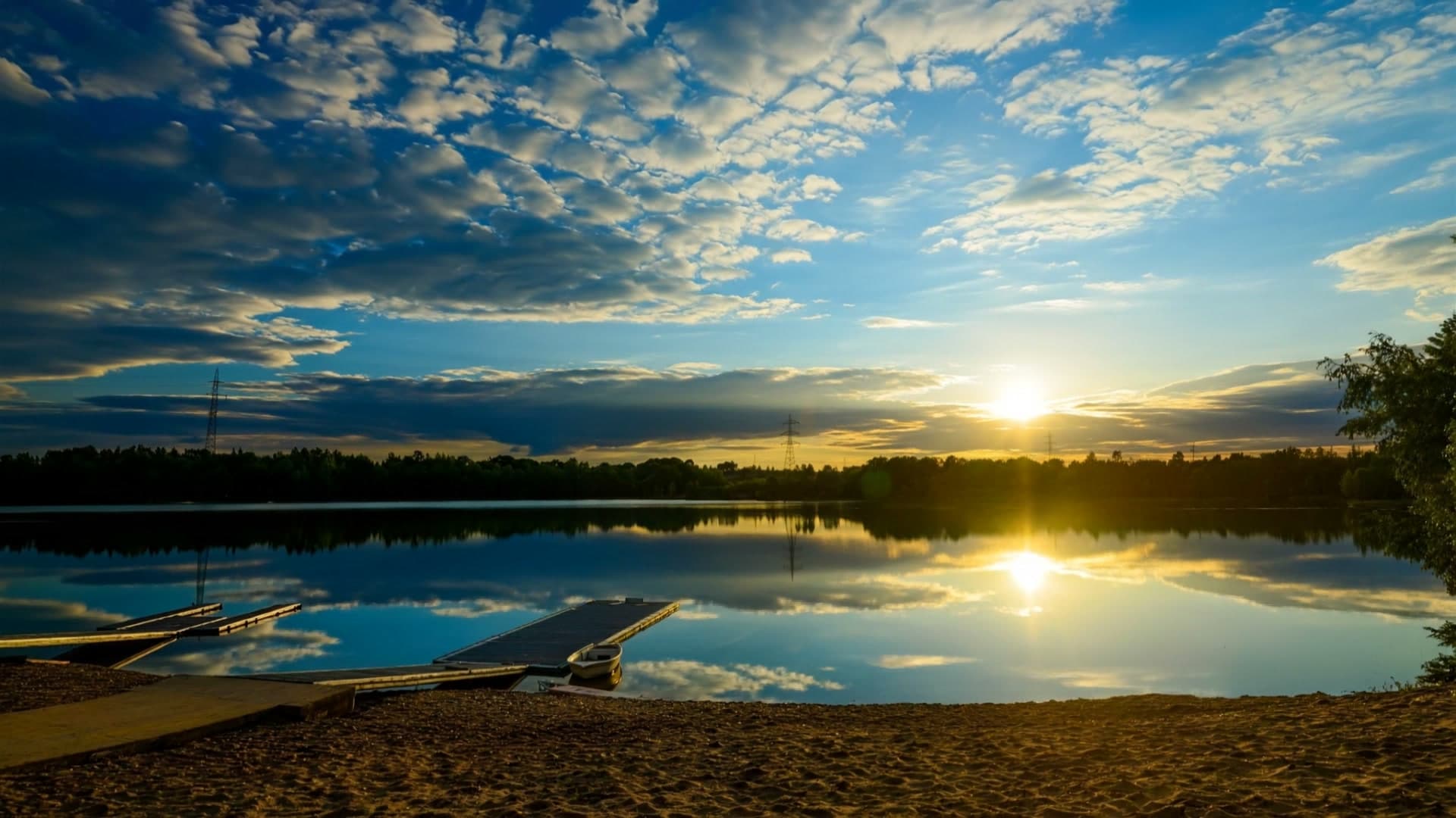 Sonnenuntergang über dem Sandhofsee: Die Sonne spiegelt sich im ruhigen Wasser, im Vordergrund Sandstrand und schwimmende Holzstege mit einem kleinen Ruderboot, am Horizont Baumlinie und Strommasten.