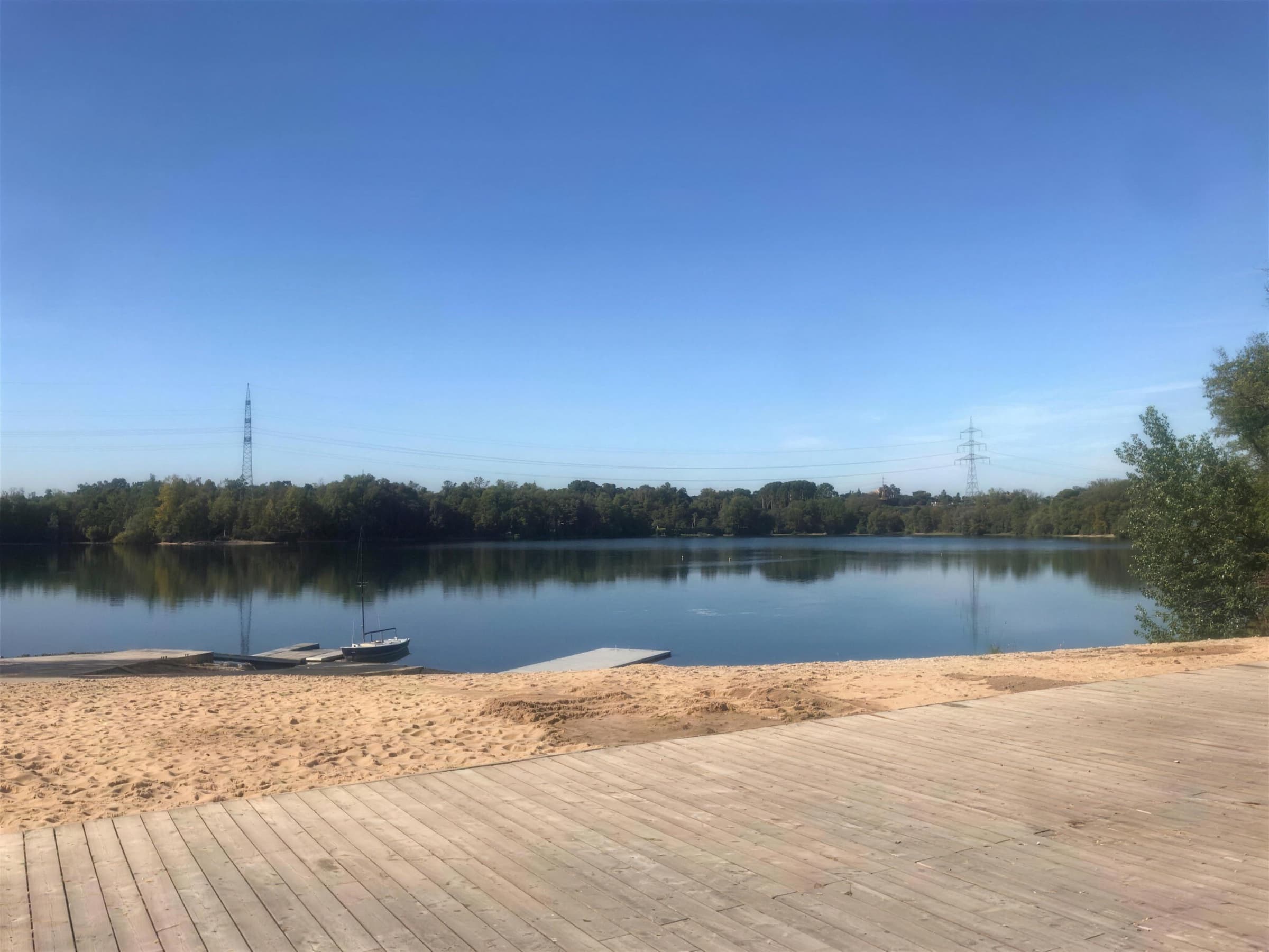 Blick von der Holzterrasse über den hellen Sandstrand auf den Sandhofsee mit Stegen und einem kleinen Segelboot, am anderen Ufer Bäume und zwei Strommasten unter blauem Himmel.