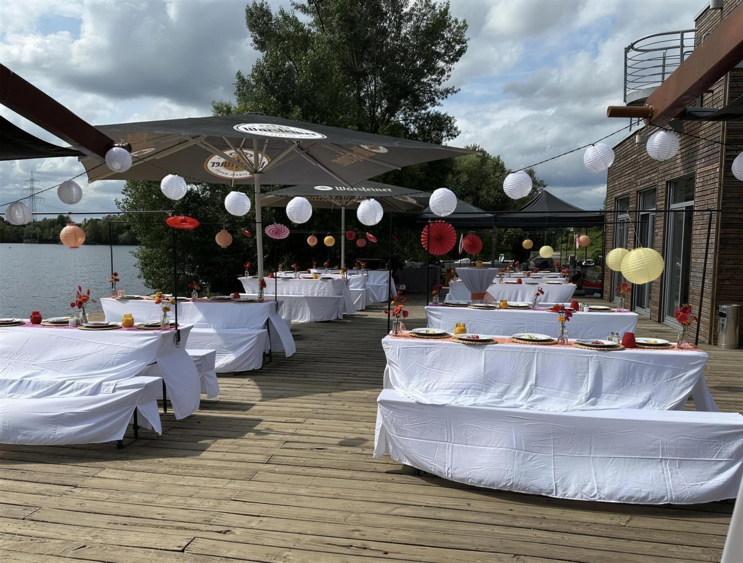 Gedeckte Tafeln mit weißen Hussen auf der Holzterrasse am See, darüber Sonnenschirme, weiße und gelbe Papierlampions und rote Papierfächer.