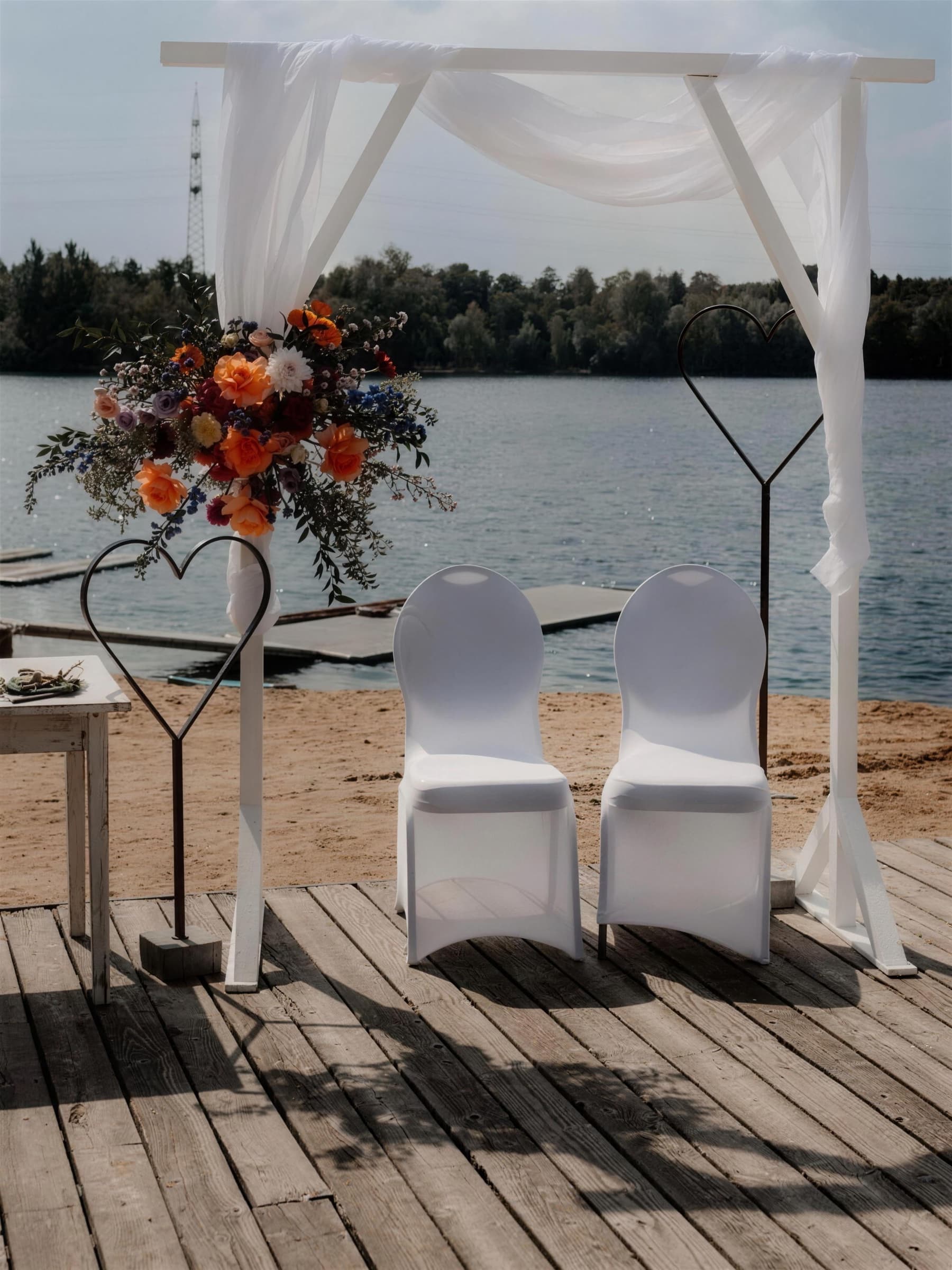 Weißer Holztraubogen mit Tüll und einem Strauß aus orangefarbenen Rosen auf dem Holzdeck am Strand, davor zwei weiß bezogene Stühle und zwei Herzen aus Metall, dahinter der See.