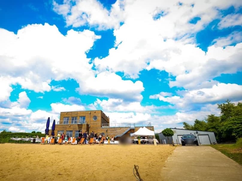 Blick über den Sandstrand auf ein zweigeschossiges Gebäude mit Holzfassade, davor Gäste, Liegestühle und ein weißes Zelt; rechts eine Zufahrt mit parkendem Auto vor einer Halle, darüber blauer Himmel mit großen Wolken.